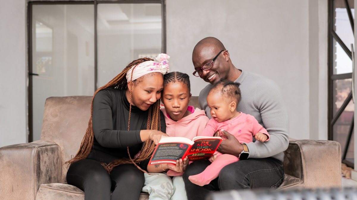 Loving black family reading a book at home on the couch, mom wearing black and kids wearing pink.