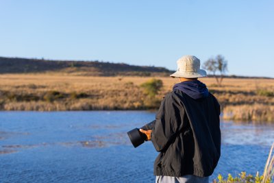 Black man standing by a river holding a camera.