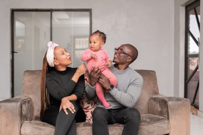 Black family smiles at home. Dad, Mom in black, and their 1-year-old daughter in pink.