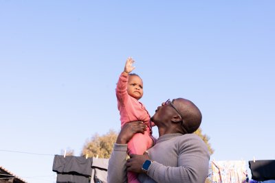 Black father playing outside with daughter wearing pink and clothes in the background with blue sky.