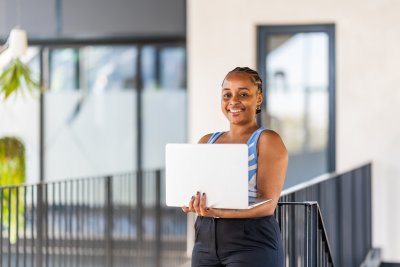 Portrait of a cheerful businesswoman in modern office and looking at camera. Smiling African executive using laptop while working from office.