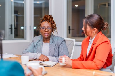 Mature black woman sitting at desk wearing glasses speaking to a candidate at an interview at the office for employment.