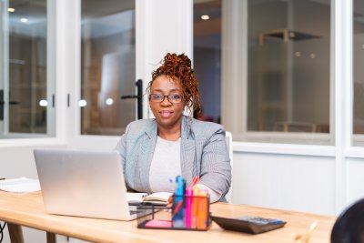 Mature African black woman sitting at desk with laptop and wearing glasses in a co working space.